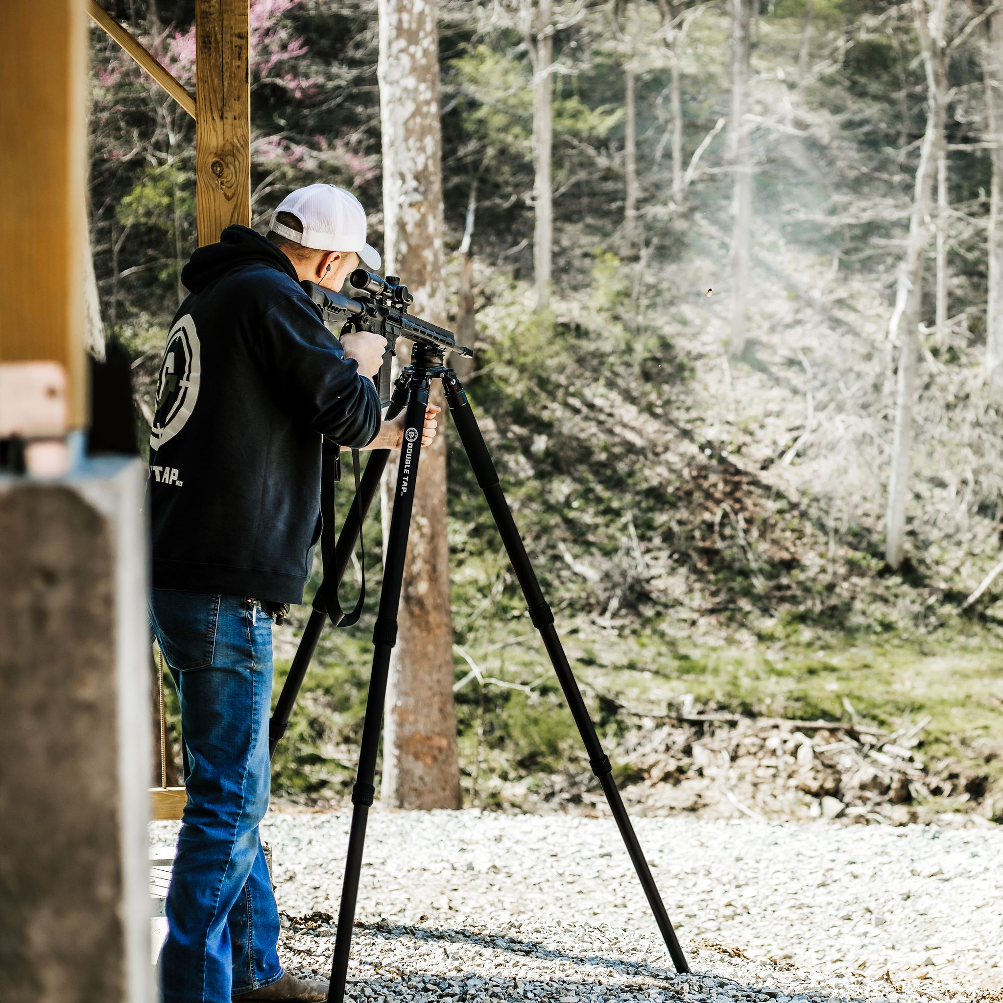 A man standing at a shooting tripod with a rifle mounted, aiming for a target in the distance