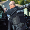 A police officer in a black tactical uniform and caliber armor patrol vest stands by an open car door, aiming a handgun through the window. The vest has the word "POLICE" printed in white on the back.