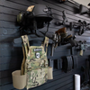 Loaded black plastic plate carrier and helmet hanging system for duty or  tactical gear mounted to Assault Wall slat wall with guns and accessories organized in the blurred background
