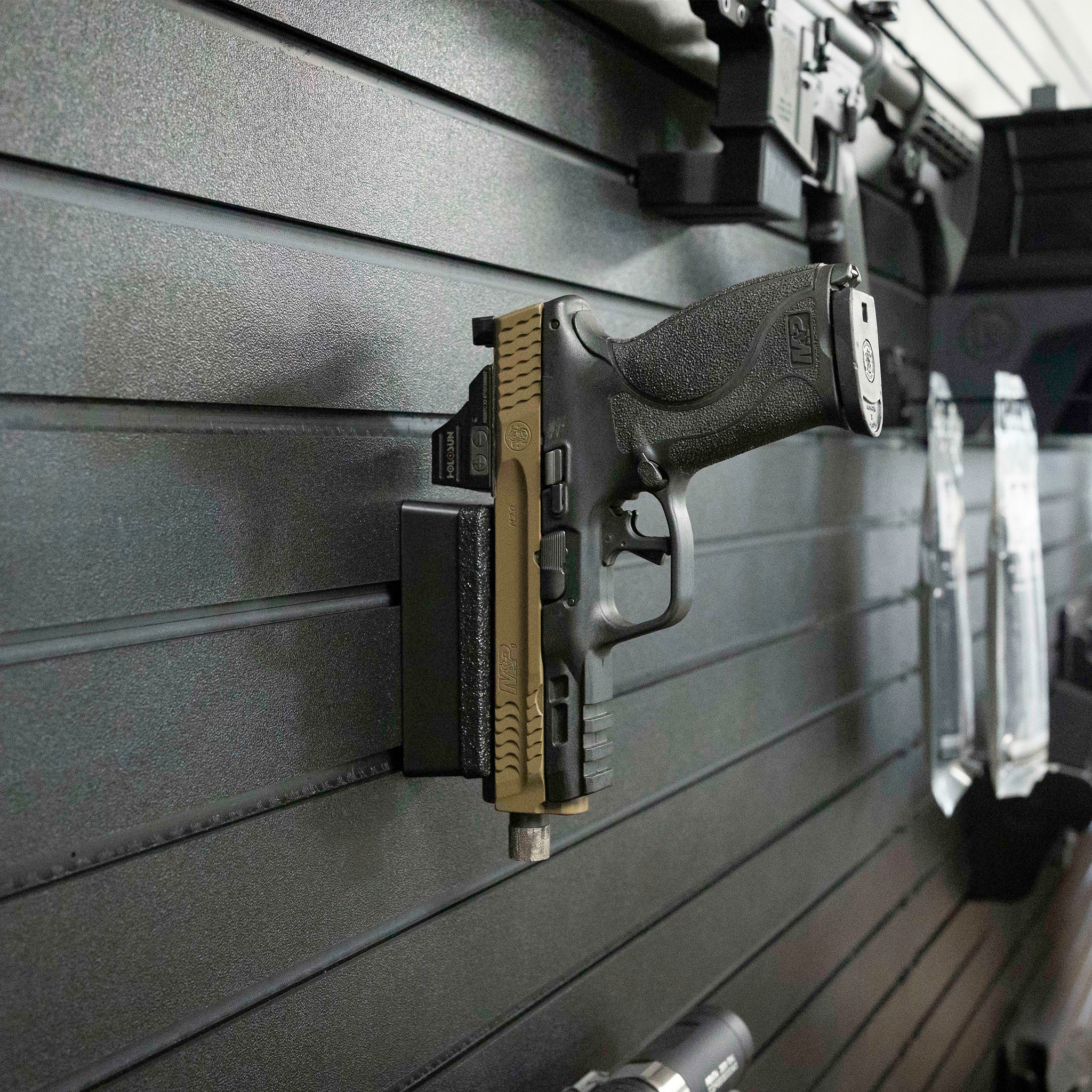 Assault Wall Magnetic gun mount shown with pistol mounted vertically by built in magnets, mount has been slid into position on a black Assault Wall slat panel