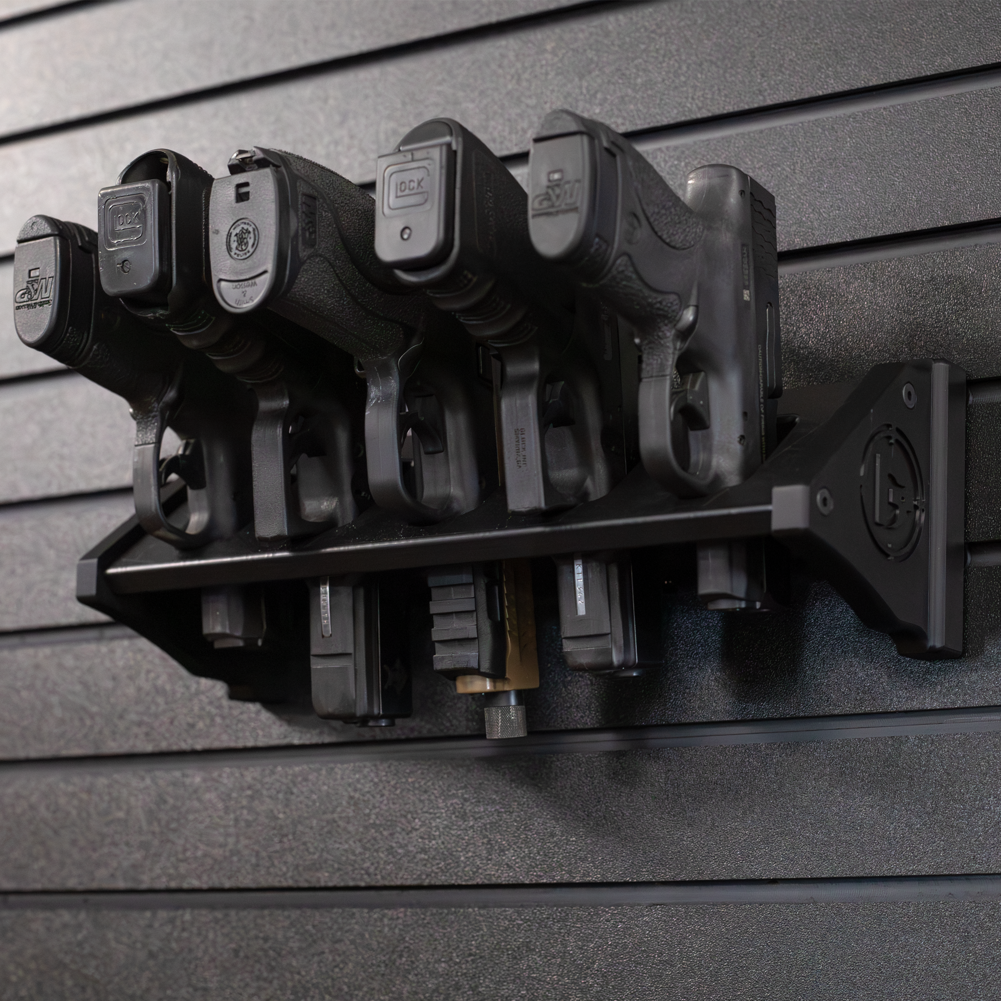 Black plastic five pistol storage mount loaded with smith and weston and glock pistols , all five pistols are resting in equally spaced slots, mount is hanging from black tactical slat wall