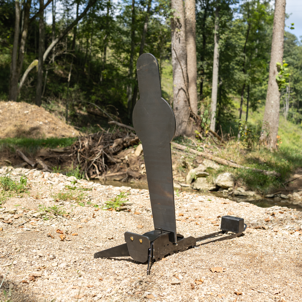 Angled view of AR500 fall popper with black metal base & inluded rubber bumper for forward or rear falling with a 42 inch uspsa popper target in wooded surroundings